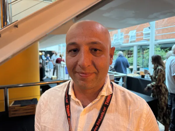 A bald man in a white shirt and lanyard stands indoors, with people and large windows in the background.