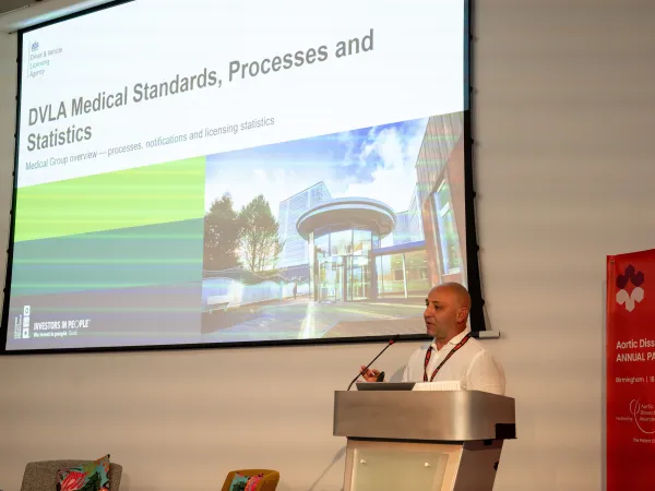 A man speaks at a podium in front of a projected slide titled 'DVLA Medical Standards, Processes and Statistics', with a red event banner beside him.